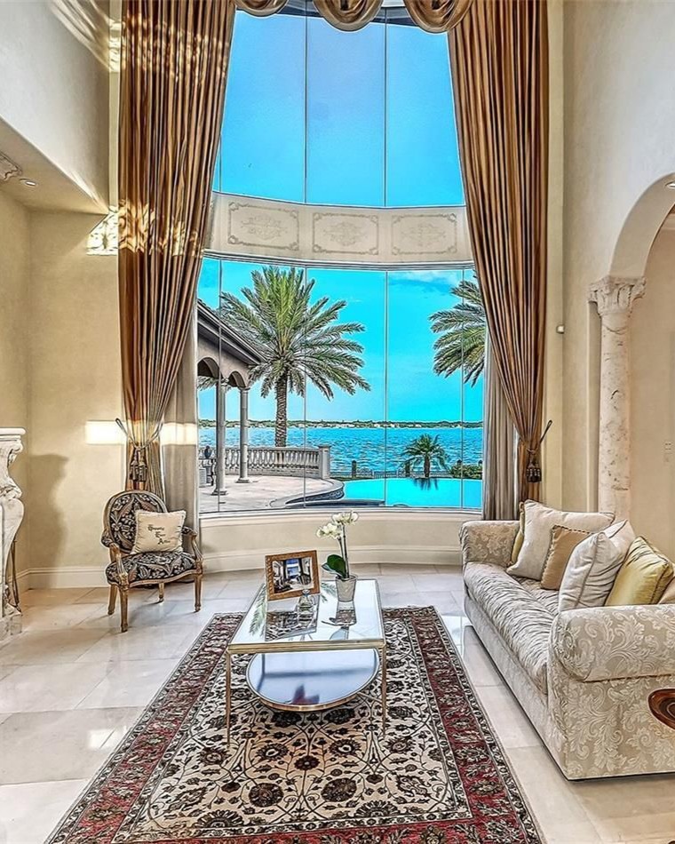 Opulent waterfront estate living room with floor-to-ceiling windows overlooking Sarasota Bay and palm-lined infinity pool.