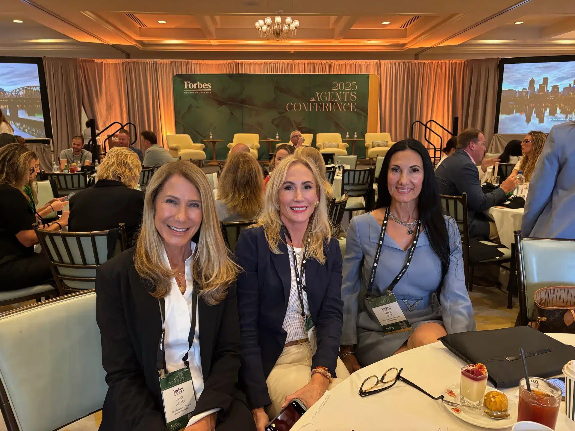 Attendees seated at the 2025 Forbes Global Properties Agents Conference, featuring three women smiling at a round table with conference badges, notebooks, and refreshments, with the stage backdrop reading “Forbes Global Properties 2025 Agents Conference.”