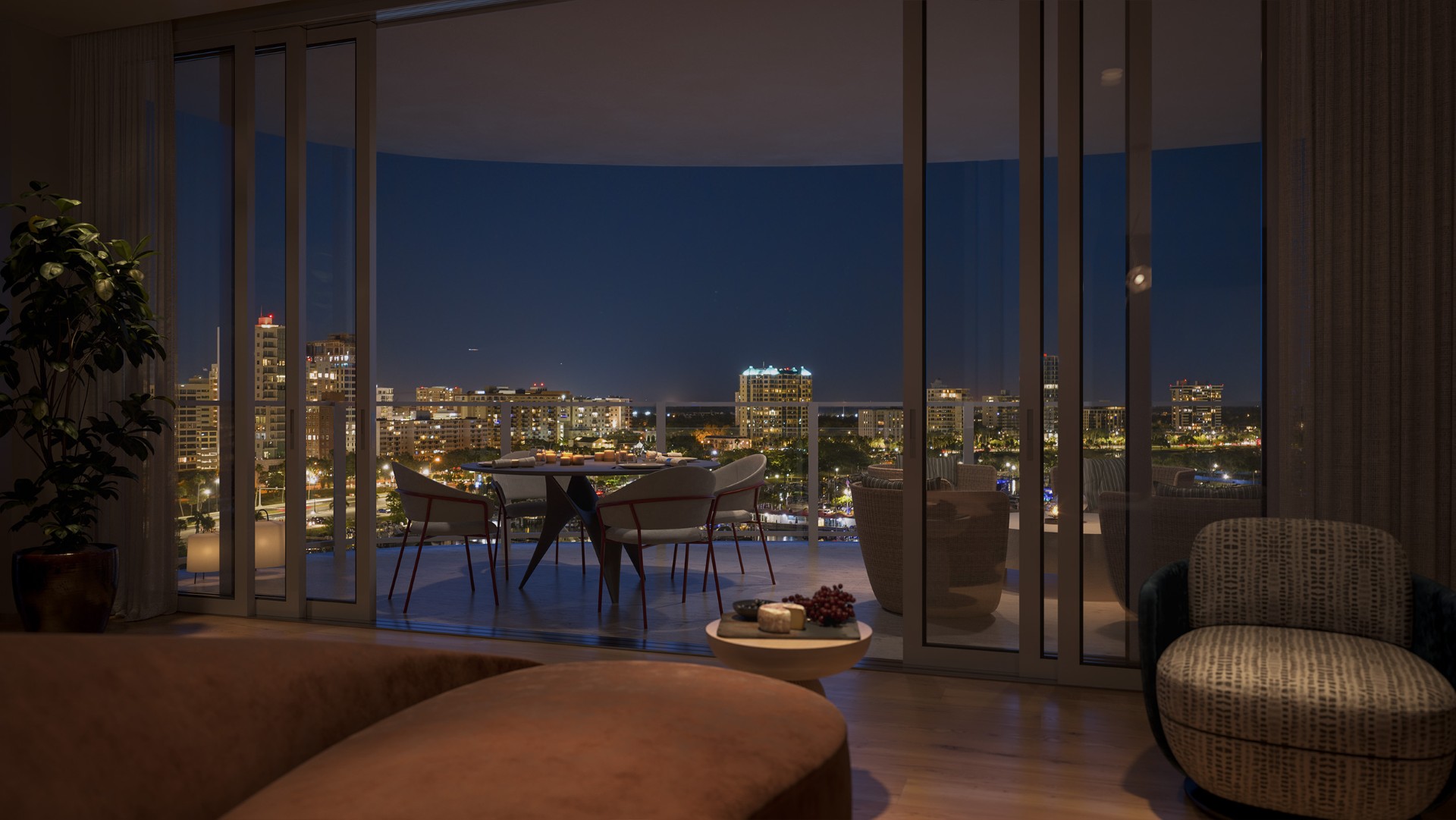 Evening view from a luxury residence at Amara on Sarasota Bay showing a private terrace, interior seating area, and illuminated downtown Sarasota skyline beyond floor-to-ceiling windows.