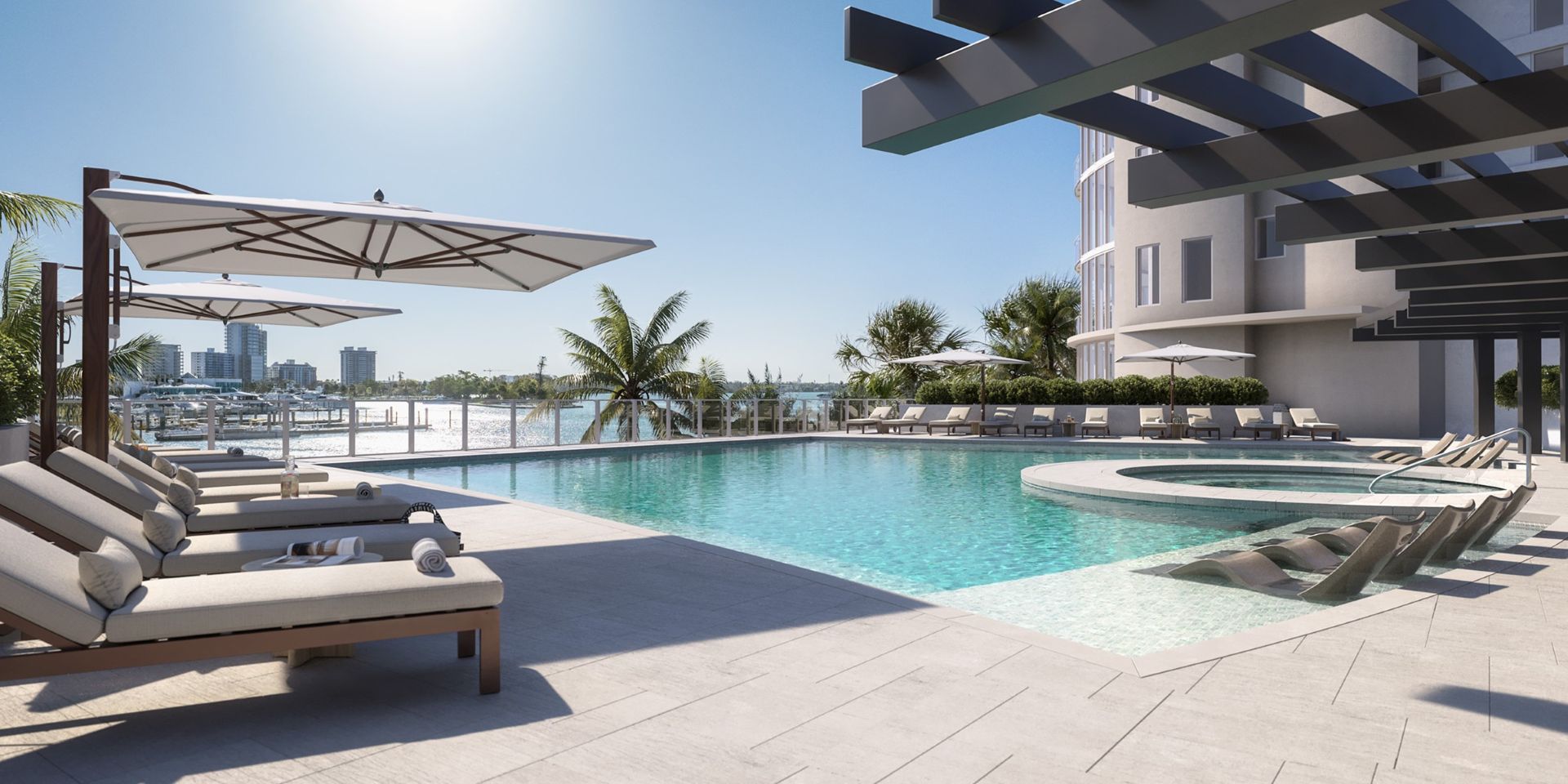 Elevated bayfront pool at Amara on Sarasota Bay featuring lounge chairs, in-water sun shelves, palm trees, and sweeping marina views under a clear blue sky.