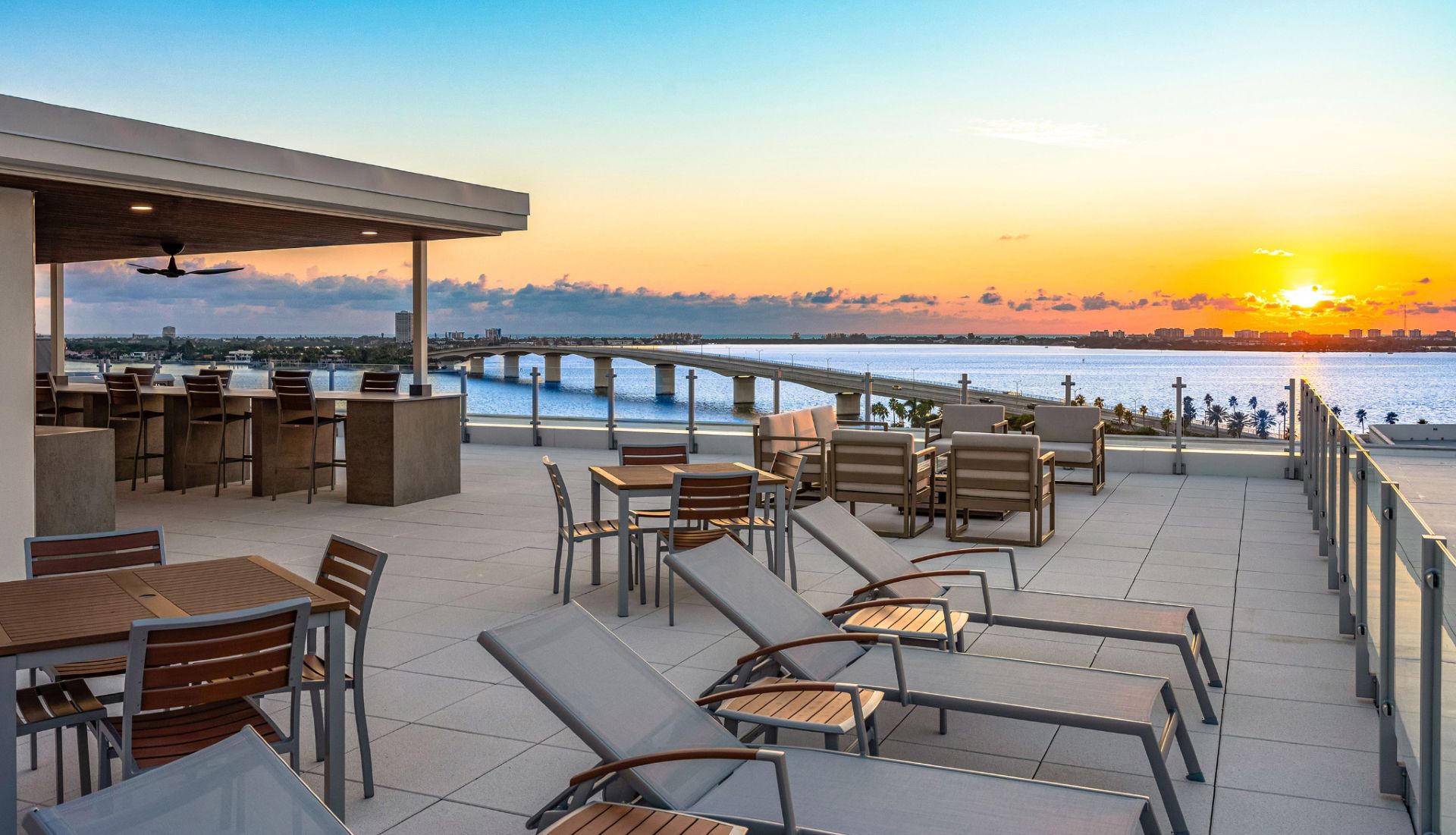 Luxury rooftop terrace featuring outdoor dining, lounge chairs, and sweeping sunset views of Sarasota Bay and the Ringling Bridge