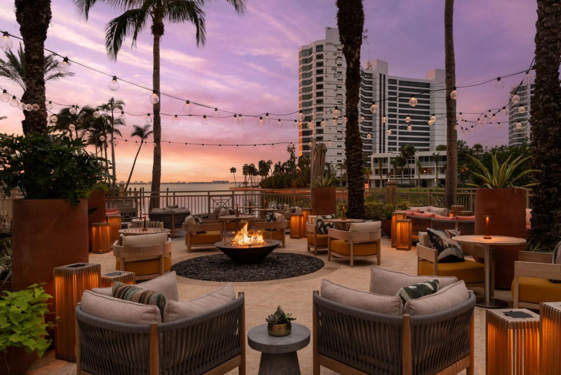 Luxury outdoor lounge with fire pit and waterfront views at The Ritz-Carlton Sarasota during sunset, featuring seating, palm trees, and ambient lighting