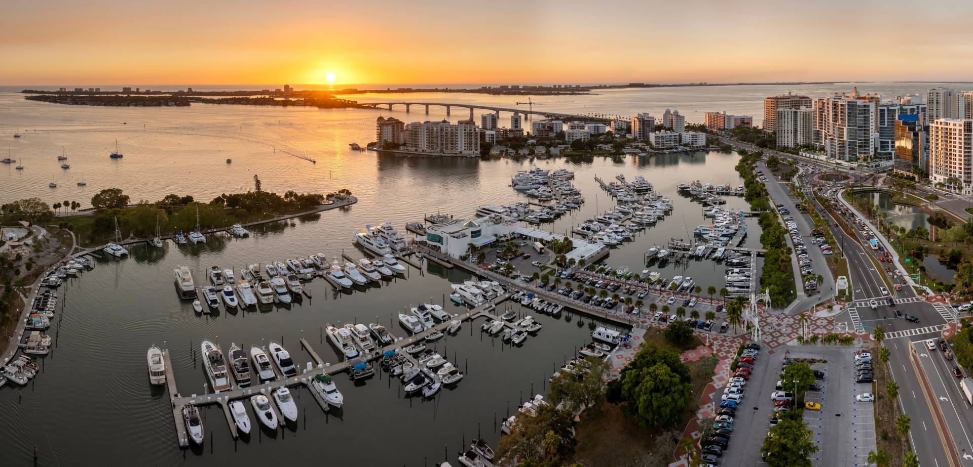 Aerial view of downtown Sarasota, Florida, at sunset featuring Sarasota Bay, the marina filled with yachts, the Ringling Bridge, waterfront high rises, and the Gulf Coast skyline.